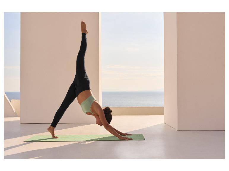 Femme faisant du yoga en extérieur avec vue sur la mer, portant un legging noir et un top de sport vert menthe.