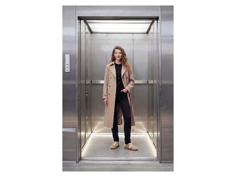 Femme en trench-coat et tenue noire debout dans un ascenseur.