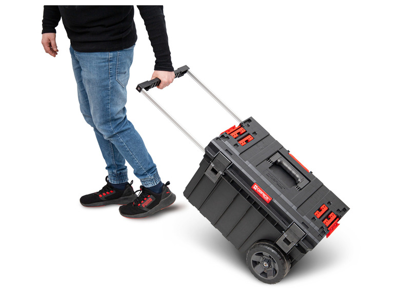 Une personne pousse une boîte à outils Qbrick System noire à roulettes.