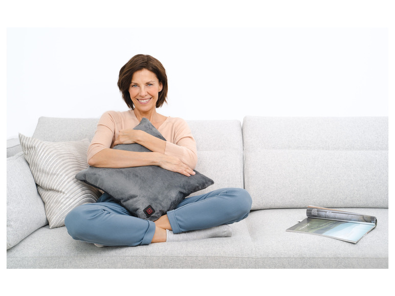 Une femme assise sur un canapé gris avec un coussin chauffant.