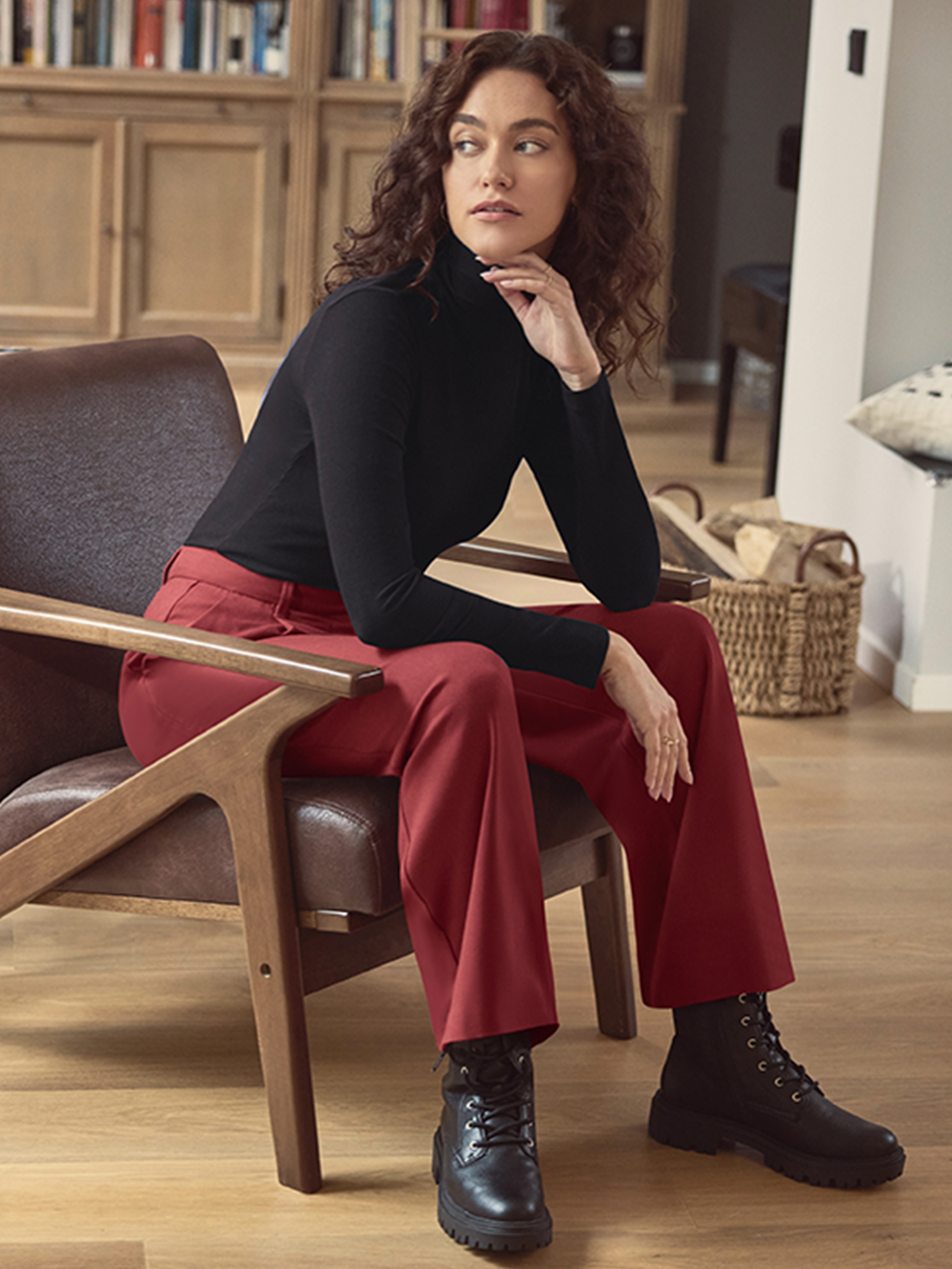 Femme en col roulé noir, pantalon rouge et bottes noires assise sur une chaise.