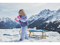 Fille en veste de ski, pantalon et gants roses, jouant avec la neige à côté d'une luge.