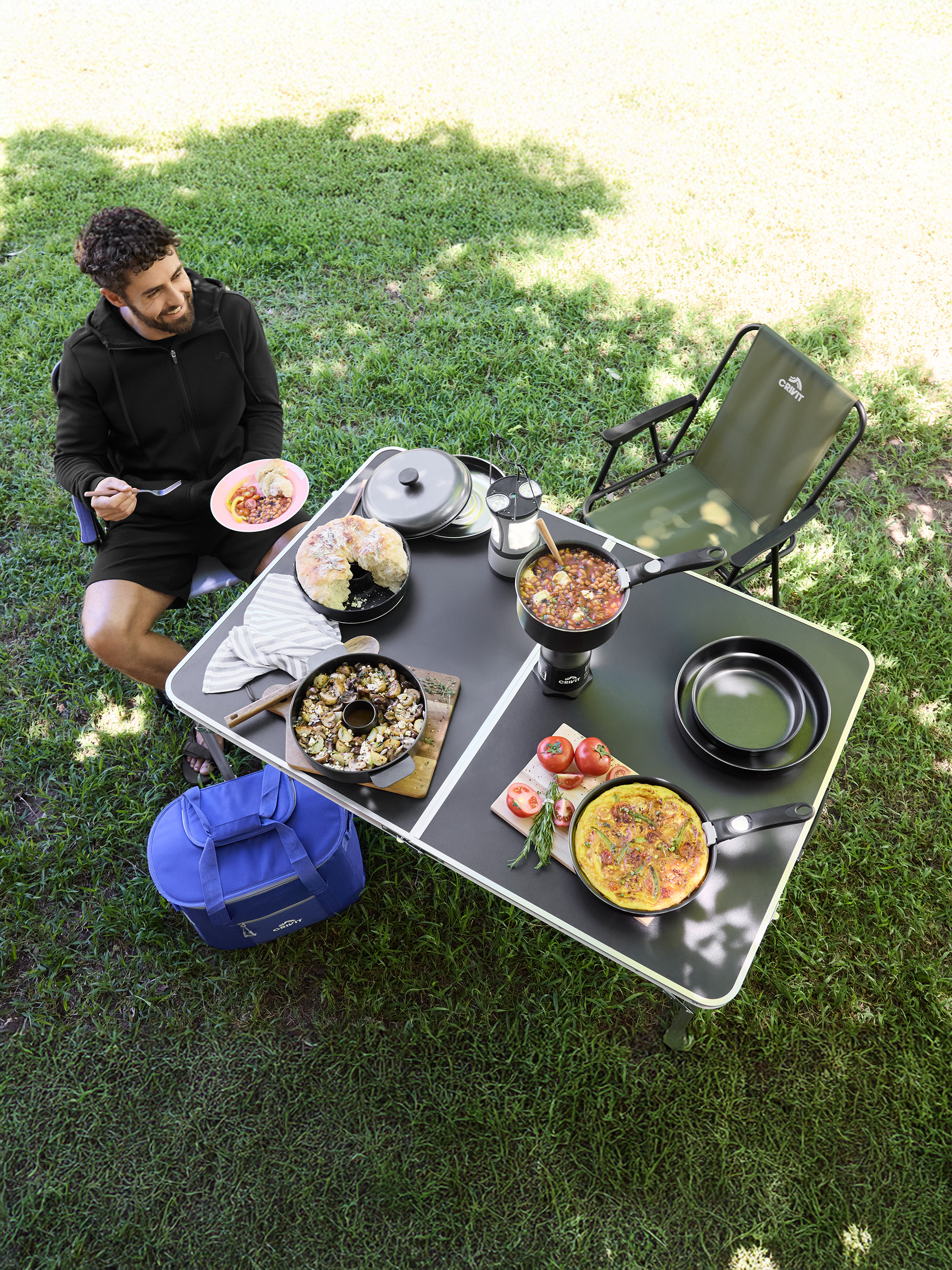 Homme savourant un repas à une table de camping avec divers plats et ustensiles de cuisine.