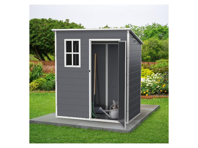 Abri de jardin gris avec porte ouverte, montrant des outils de jardinage, posé sur une base en béton sur une pelouse.