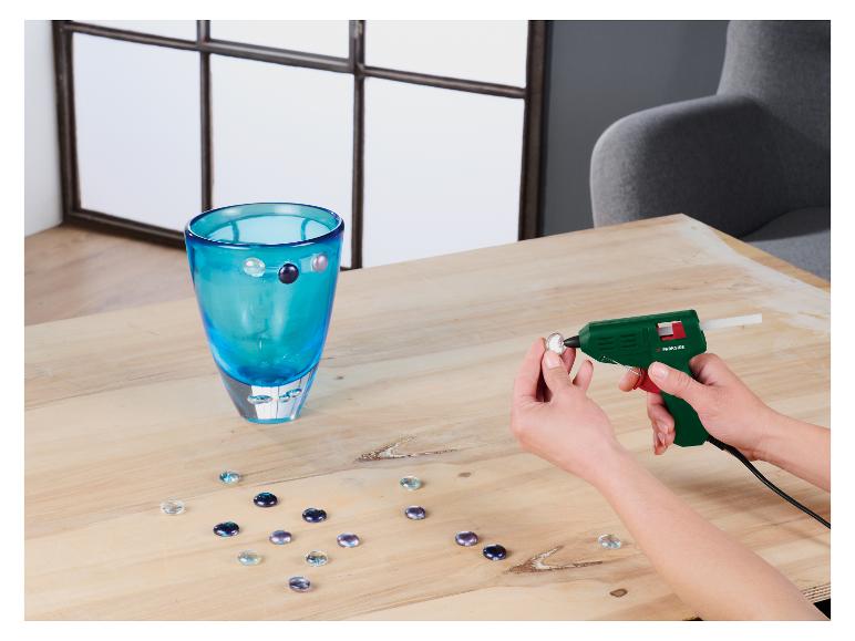 Une personne décore un vase turquoise avec des pierres de verre à l'aide d'un pistolet à colle.