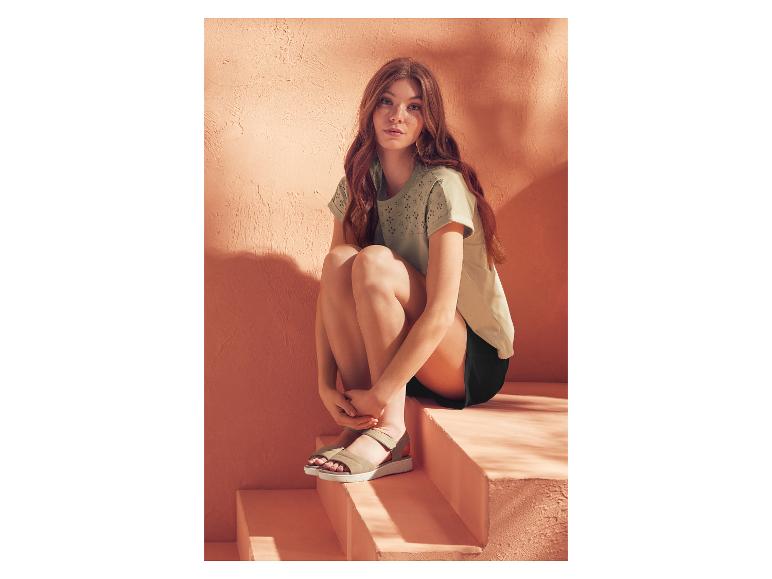 Femme en t-shirt vert clair et short noir assise sur des marches et portant des sandales beige clair.