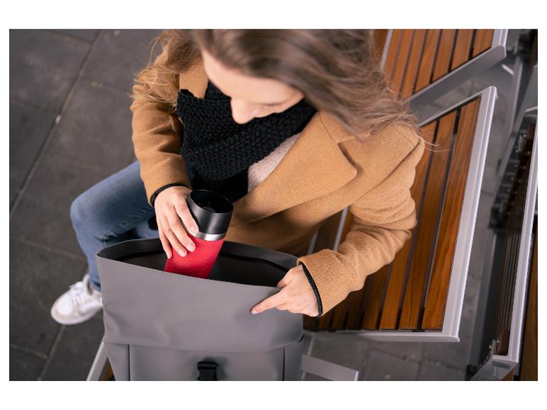 Femme mettant une tasse de voyage rouge dans un sac à dos gris sur un banc.