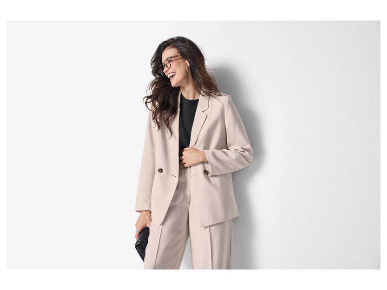 Femme en blazer et pantalon clairs, t-shirt noir et lunettes, souriant de côté.