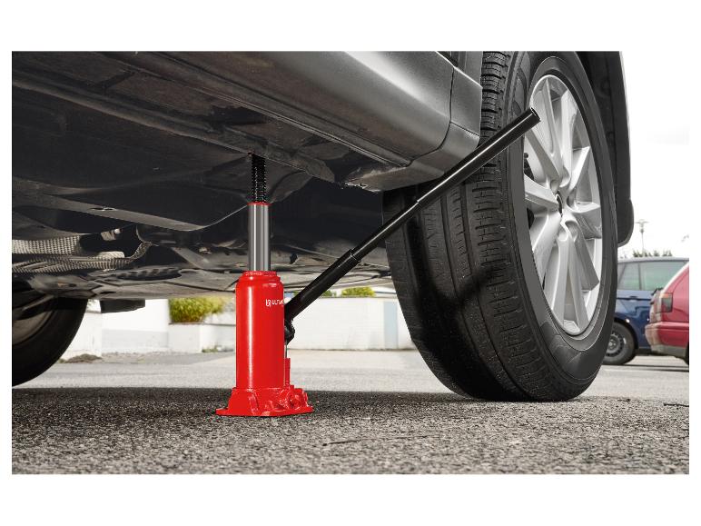 Cric hydraulique rouge sous une voiture grise, la soulevant légèrement du sol.