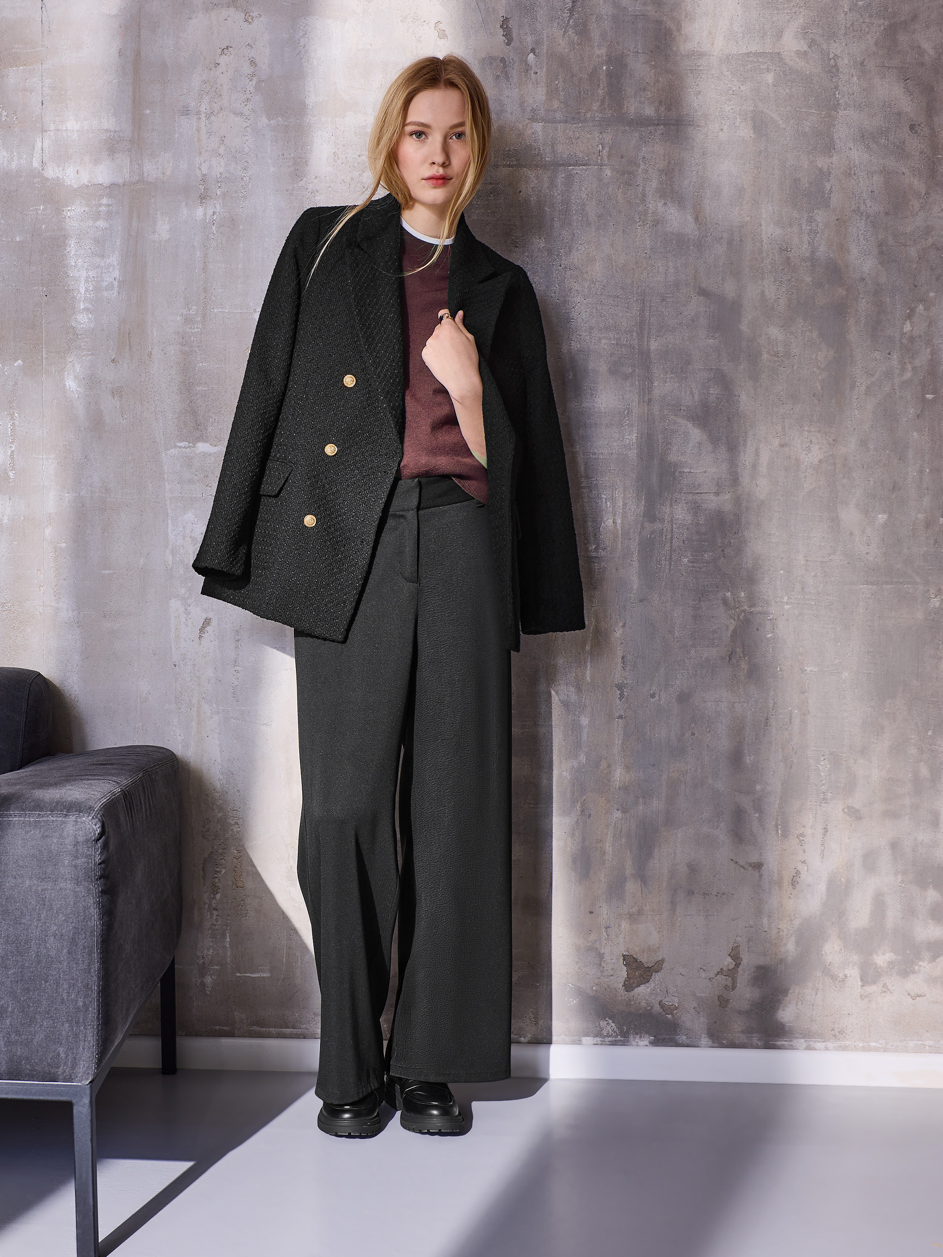 Femme en blazer noir, pull bordeaux et pantalon large noir, posant devant un mur texturé.