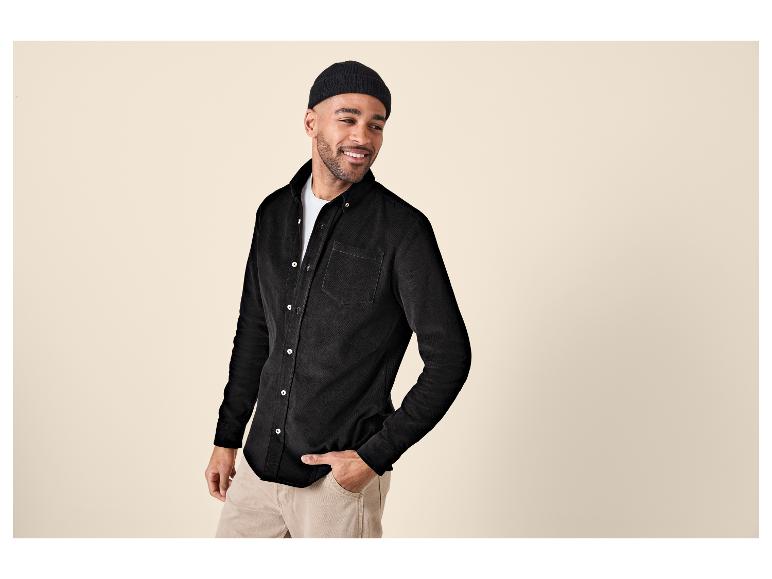 Homme en chemise en velours côtelé noire et pantalon beige, souriant et regardant de côté.