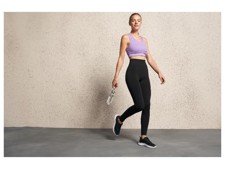 Une femme en leggings noirs et un soutien-gorge de sport lila pose avec une bouteille d'eau.