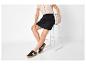 Femme en chemise beige, jupe noire et baskets noires assise sur une chaise.