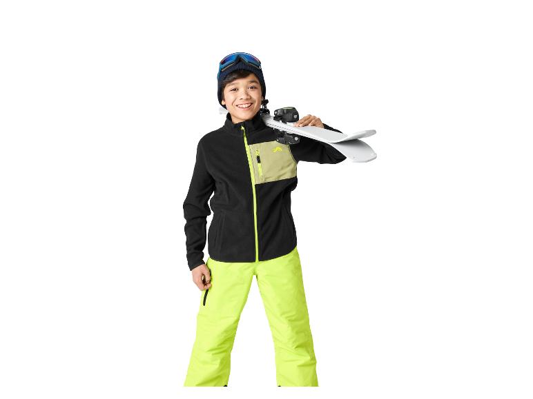 Un jeune garçon en veste polaire noire et pantalon de ski jaune fluo tient des skis sur son épaule.