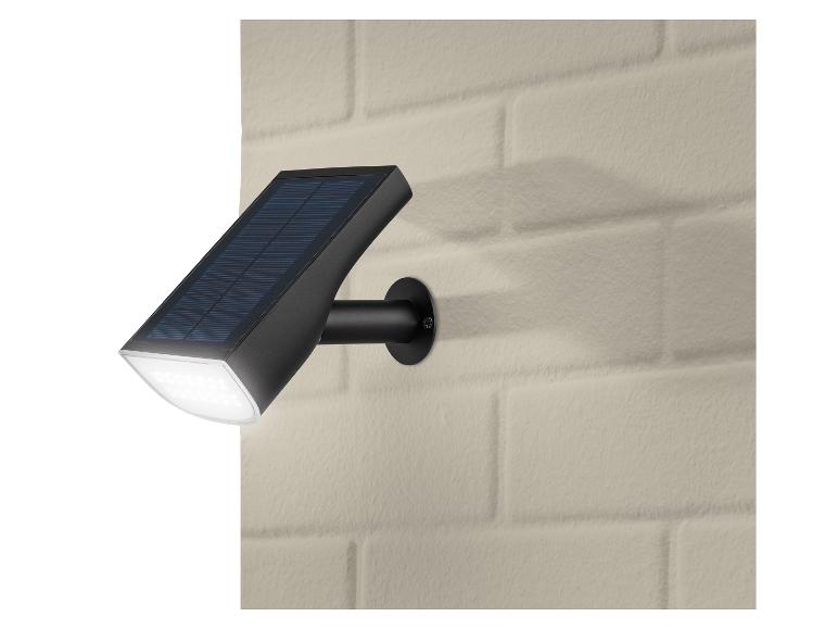 Lampe solaire LED noire avec panneau solaire, montée sur un mur de briques beige.