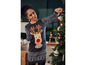 Une femme portant un pull de Noël avec un renne accroche des décorations de Noël à un sapin.