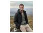 Homme en veste noire et pull vert assis sur un rocher dans un paysage montagneux.
