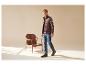 Homme en gilet marron, chemise à carreaux et jean, à côté d'une chaise en bois.