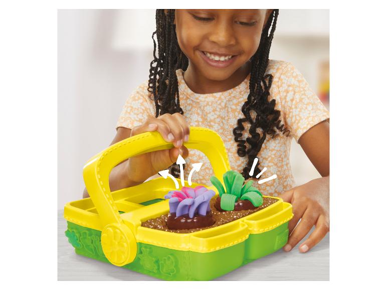 Enfant souriant jouant avec un panier de jardinage Play-Doh avec des fleurs et des légumes en pâte à modeler.