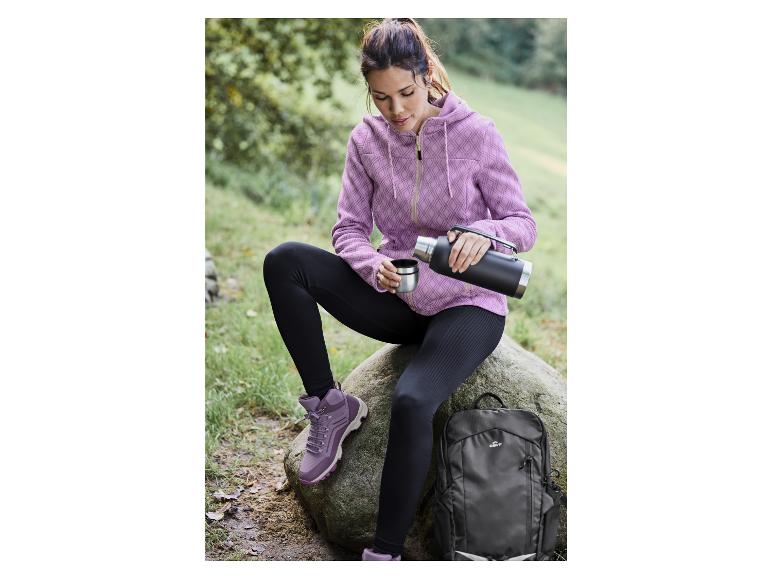 Une femme en sweat-shirt rose, leggings noirs et chaussures de randonnée avec une thermos et un sac à dos Crivit.