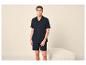 Homme en chemise et short bleu marine, debout devant un pouf blanc.