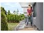 Femme nettoyant une terrasse avec un nettoyeur haute pression, avec des buissons et arbres verts en arrière-plan.