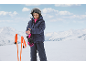 Enfant en veste de ski, pantalon de ski, casque et gants, ajustant un gant avec des bâtons de ski à proximité.