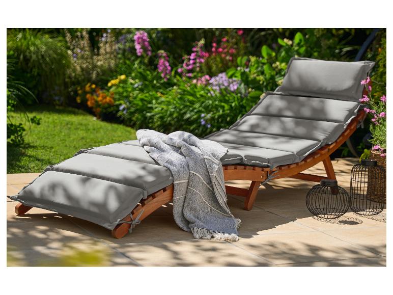 Chaise longue de jardin en bois avec coussins gris et couverture sur une terrasse fleurie.