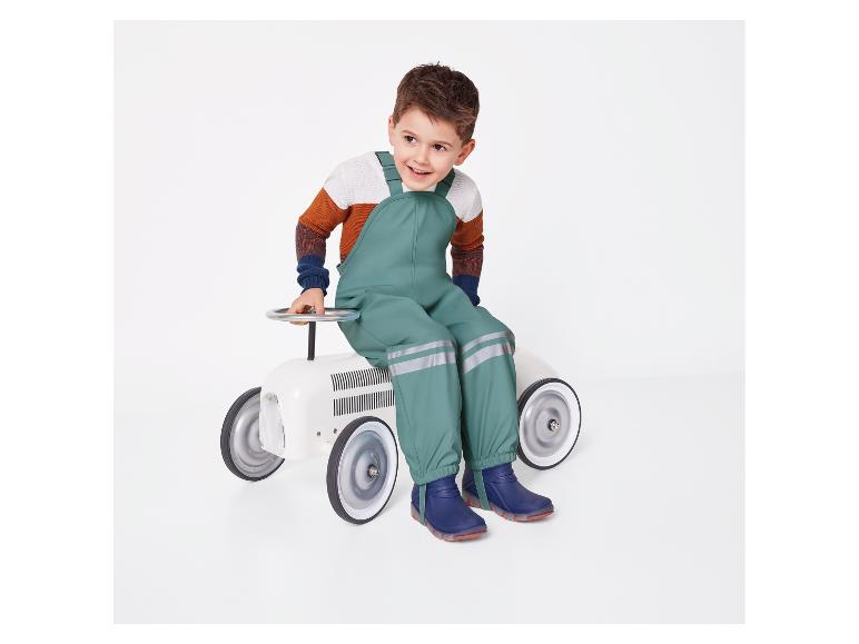 Un enfant en salopette verte et bottes bleues est assis sur une voiture jouet blanche.
