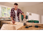 Homme vissant une poutre en bois avec une perceuse sans fil, avec une boîte à outils et un chargeur sur la table.