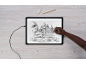 Une personne dessine un moulin au bord d'une rivière sur une tablette lumineuse, avec un stylo, une gomme et un crayon.