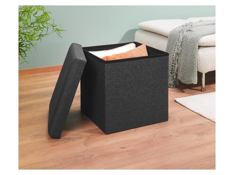 Un pouf de rangement en tissu gris foncé avec un dessus rembourré est ouvert sur un plancher de bois clair.