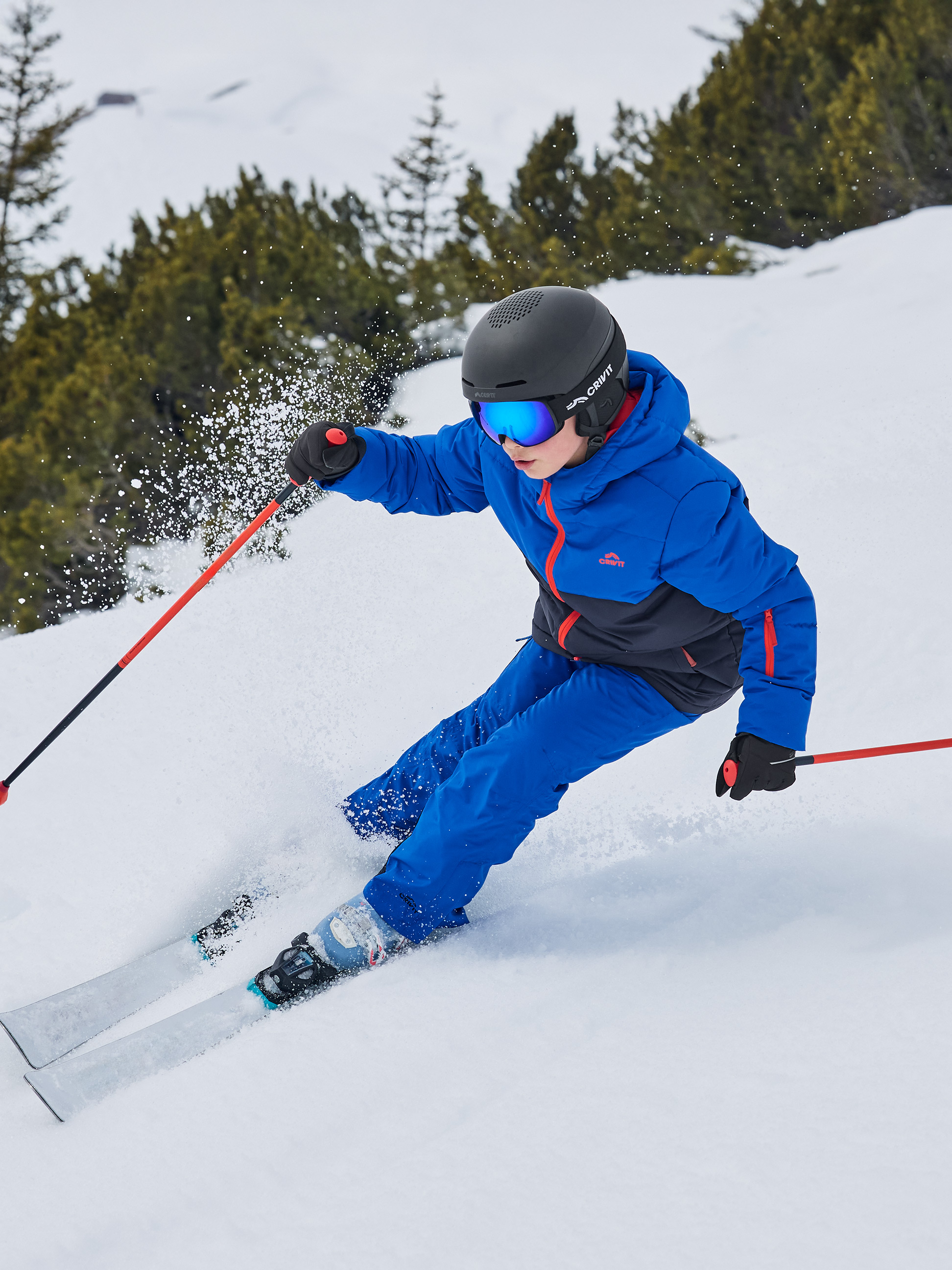 Enfant en veste et pantalon de ski bleus, casque et lunettes, skiant sur une pente enneigée.