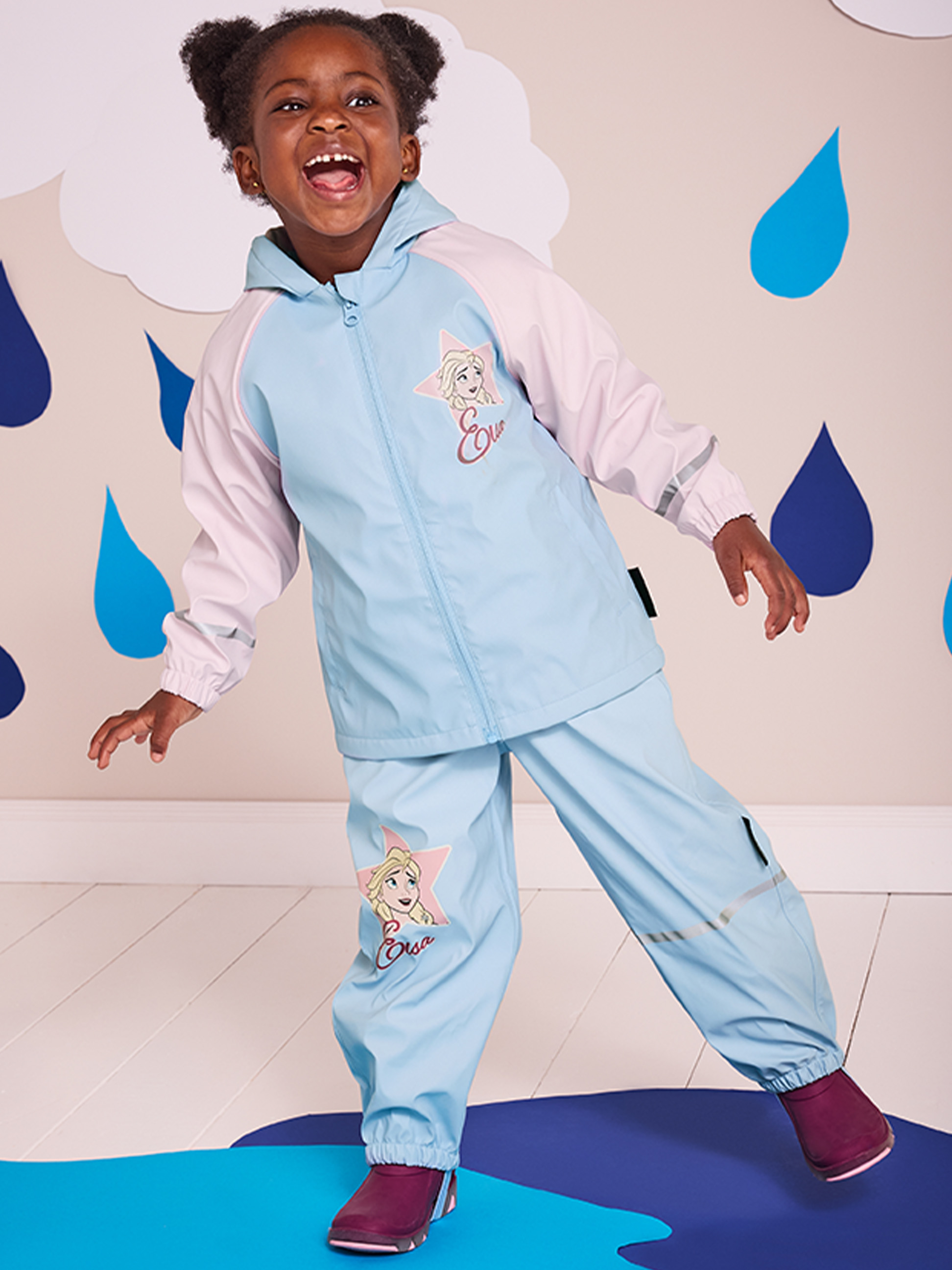Un enfant en combinaison de pluie La Reine des Neiges bleu clair et rose, souriant sur un tapis bleu.