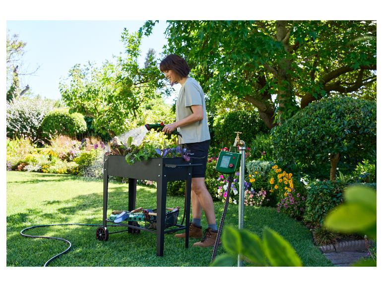 Femme arrosant des plantes dans un potager surélevé avec un tuyau, à côté d'un programmateur d'arrosage.