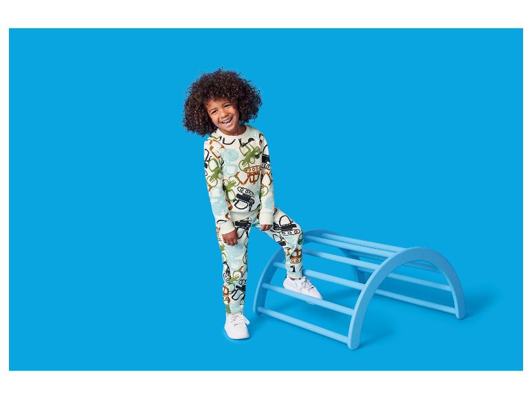 Enfant heureux en pyjama imprimé véhicules, debout sur un cadre d'escalade bleu.