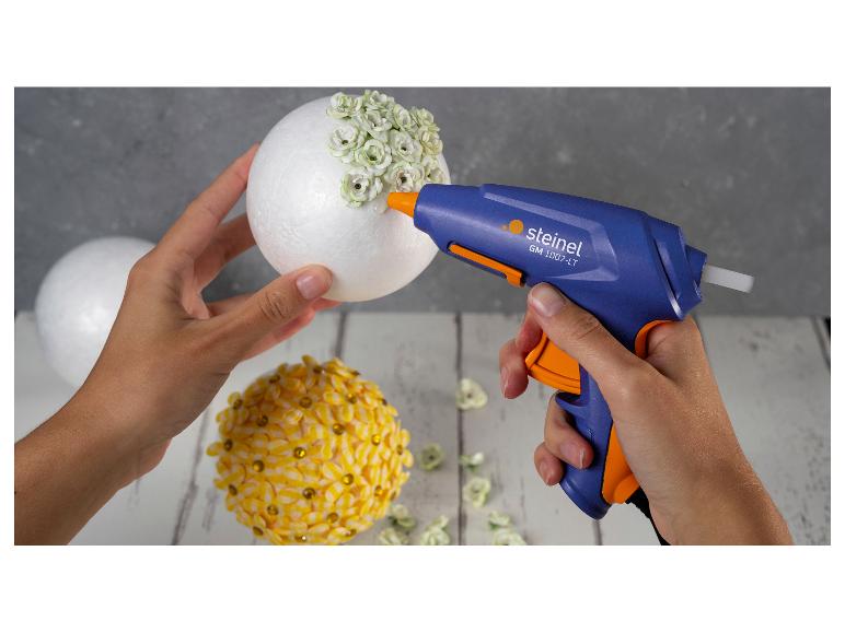 Pistolet à colle fixant des fleurs blanches sur une boule de polystyrène, une autre boule décorée de fleurs jaunes.