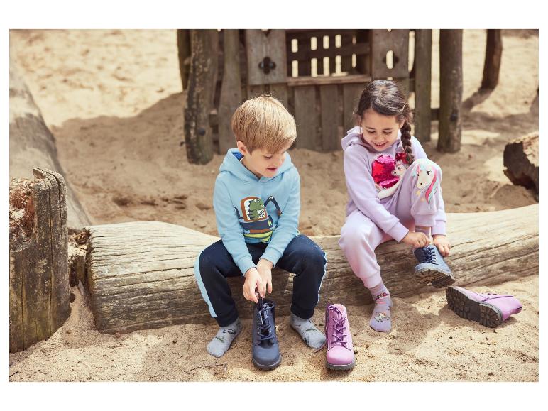 Deux enfants enfilent leurs bottes sur une bûche en bois dans une aire de jeux sablonneuse.