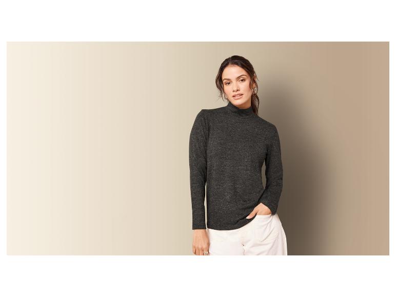 Une femme porte un pull à col roulé gris foncé et un pantalon blanc.