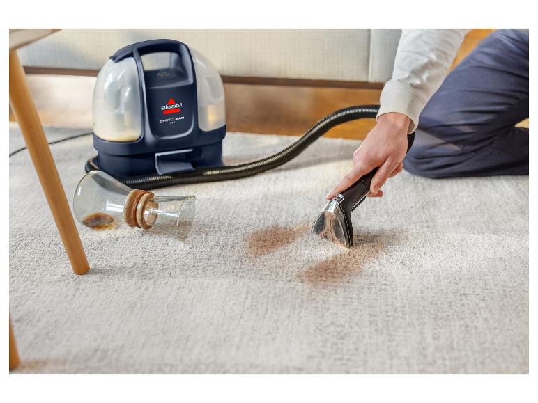 Une personne nettoie un verre de café renversé sur un tapis clair avec un nettoyeur de tapis Bissell SpotClean Mini.