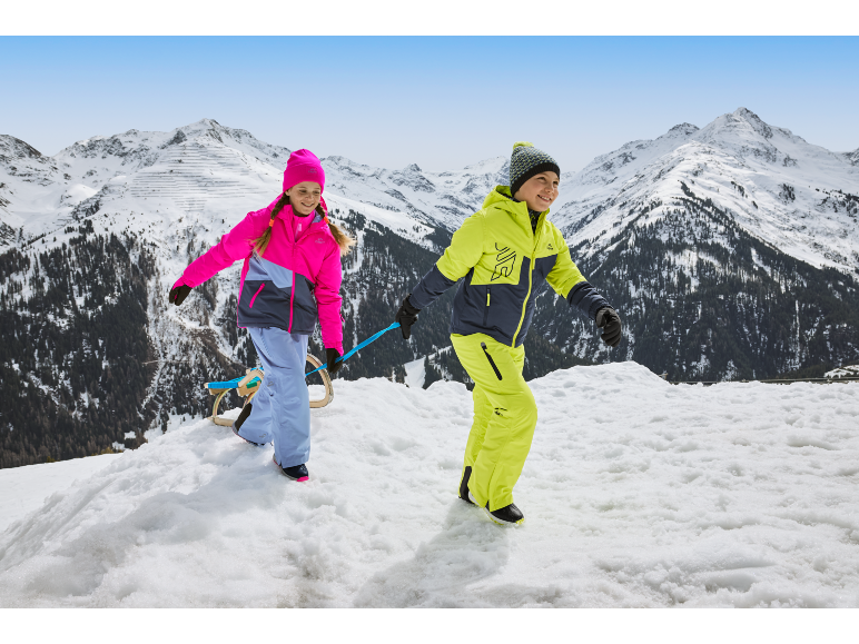 Deux enfants en combinaisons de ski et bonnets colorés, tirant une luge dans un paysage montagneux enneigé.