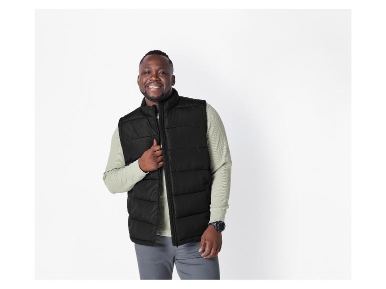 Homme souriant en gilet matelassé noir, chemise à manches longues vert clair et pantalon gris.