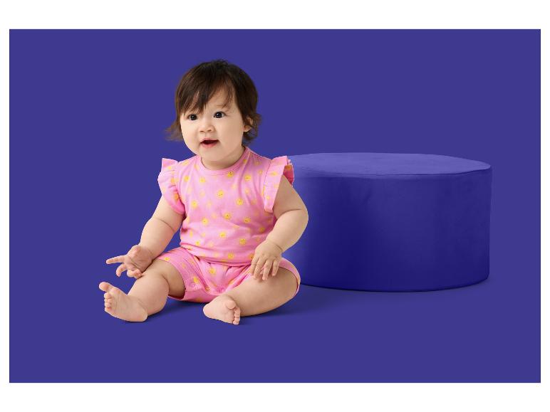 Un bébé en barboteuse rose à motif soleil assis à côté d'un pouf bleu.