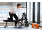 Homme s'entraînant avec un haltère sur un banc de fitness dans une salle de sport avec de grandes fenêtres.
