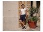 Homme en débardeur et short bleu, appuyé contre un mur décoré.