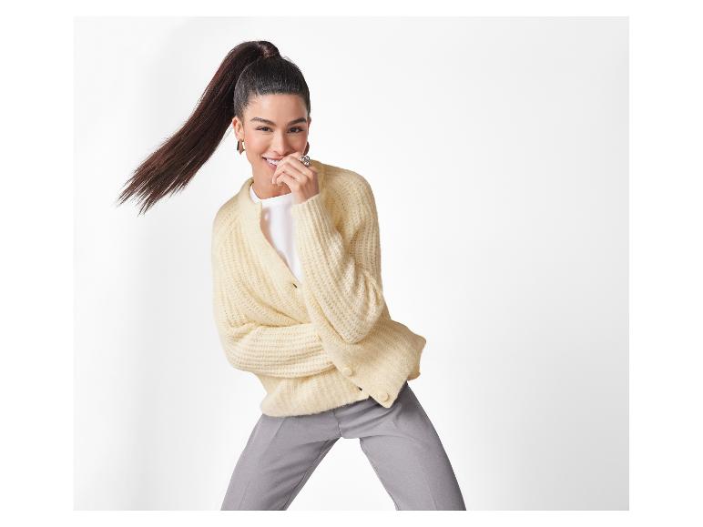 Femme souriante avec une queue de cheval, portant un cardigan crème et un pantalon gris.