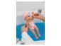 Deux mains tenant une poupée bébé en maillot de bain et bonnet roses au-dessus de l'eau bleue dans une baignoire.