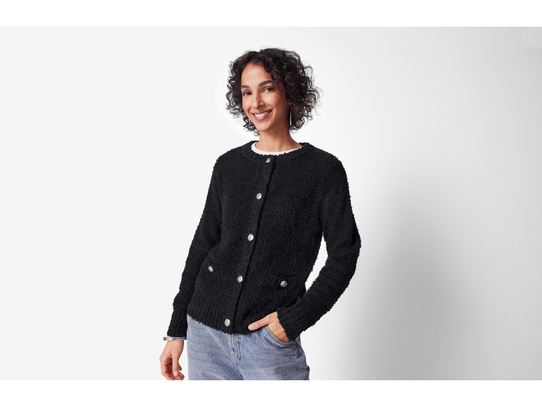 Femme souriante en cardigan noir texturé et jean, main dans la poche.