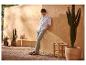 Homme en chemise blanche et pantalon gris, appuyé contre un mur en terre cuite avec cactus et plantes.