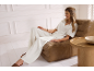 Femme en pyjama blanc se détend sur un fauteuil marron dans un salon lumineux.